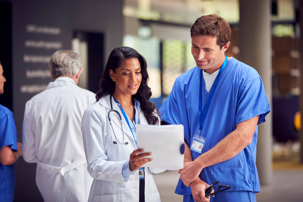 病院で正式な会議を持っているデジタルタブレット付きホワイトコートとスクラブの2人の医療スタッフ - 写真・画像