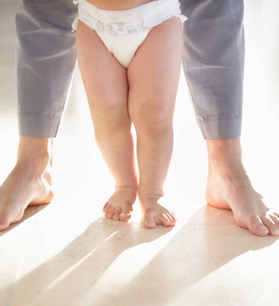 Первые шаги. Обрезанное изображение ног ребенка, идущего с родителями, поддерживающими сзади. - Фото, изображение