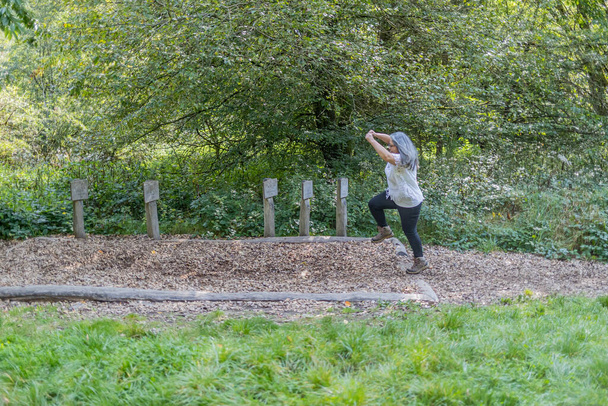 Reife Frau, die auf einem Speedtrack in einem Naturschutzgebiet in eine Erdgrube springt, sportlicher Ort an einem Wanderweg, wilde Pflanzen und Bäume im Hintergrund, sonniger Sommertag in Luxemburg. Aktives Lebenskonzept - Foto, Bild