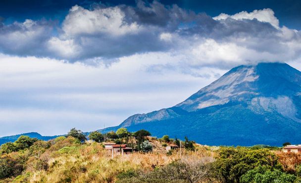 Meksykański krajobraz z domem na wzgórzu z wulkanem Colima z fumarolą w tle, słoneczny dzień z błękitnym niebem i obfite chmury w stanie Jalisco, Meksyk - Zdjęcie, obraz