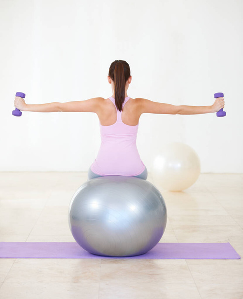 The two core components to her workout. Rearview shot of a young woman sitting on an exercise ball and doing some strengthening exercises with dumbbells. - 写真・画像