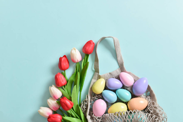 A beautiful bouquet of tulips with a string bag containing painted eggs. Banner for the Happy Easter holiday. Copy space. Flat lay, top view. - Valokuva, kuva