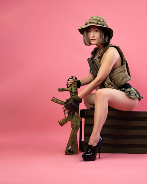 szexi ázsiai nő katonai egyenruhában automata puskával ül egy lőszeres ládán - Fotó, kép
