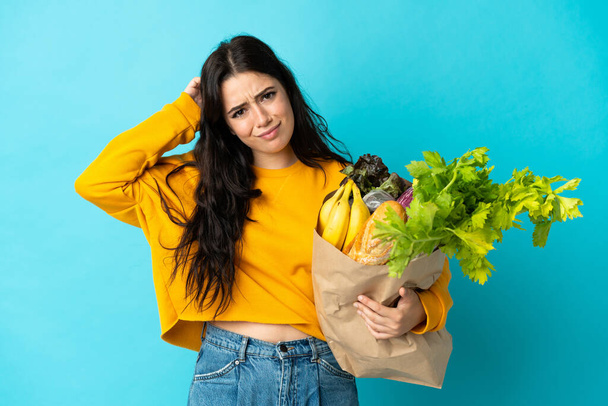 Mujer joven sosteniendo una bolsa de la compra de comestibles aislado sobre fondo azul que tiene dudas - Foto, Imagen
