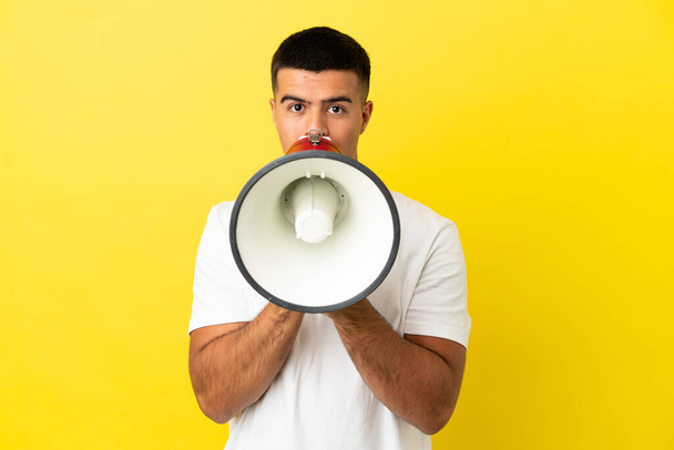 Young handsome man over isolated yellow background shouting through a megaphone to announce something - Фото, изображение