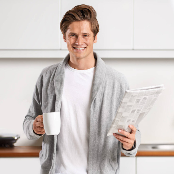 My morning ritual.... A young man standing in a kitchen drinking coffee and reading the newspaper. - Photo, image