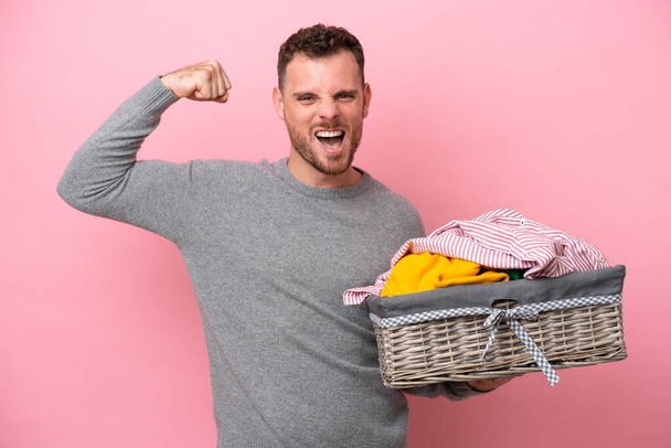 Junger brasilianischer Mann hält einen Wäschekorb auf rosa Hintergrund und macht eine starke Geste - Foto, Bild
