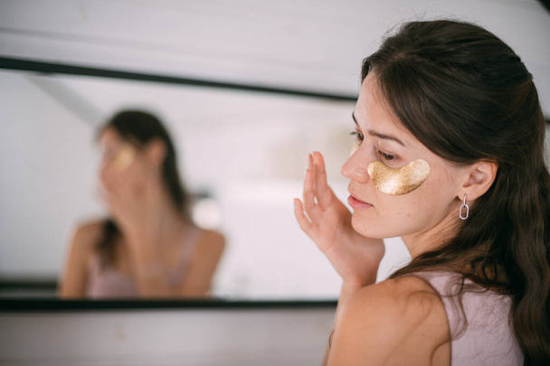 Молода жінка з плямами під очима у дзеркалі вдома. Красива брюнетка робить процедури догляду за шкірою обличчя гіалуроновими плямами біля великого дзеркала в світлій кімнаті
. - Фото, зображення