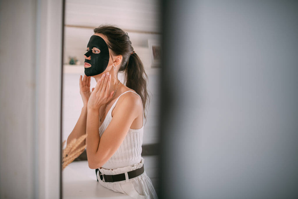 Portret van een jonge vrouw met een zwart gezichtsmasker bij de spiegel thuis. Een mooie brunette vrouw maakt gezichtsverzorging procedures met een reinigende, hydraterende masker in de buurt van de spiegel in een lichte kamer - Foto, afbeelding