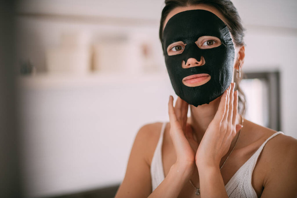 Portrait of a young woman with a black face mask at the mirror at home. A beautiful brunette woman makes facial skin care procedures with a cleansing, moisturizing mask near the mirror in a bright room - Photo, Image