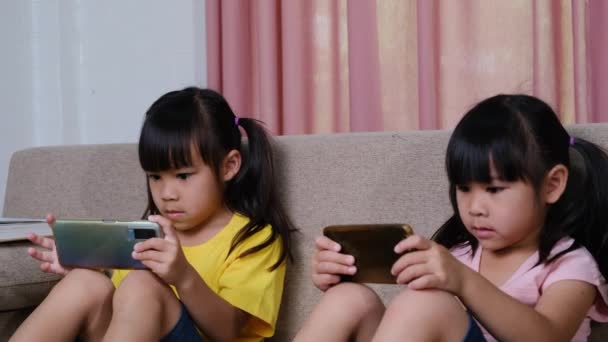 Zwei Schwestern spielen Online-Spiele auf ihren Smartphones und sitzen zu Hause auf dem Sofa. Modernes Kommunikations- und Gadget-Suchtkonzept. Zwei Kinder mit Gadgets. - Filmmaterial, Video