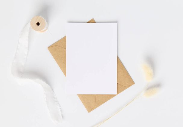 Blank πρόσκληση ευχετήρια κάρτα Mockup σε καφέ φάκελο με αποξηραμένα λαγουδάκι ουρές γρασίδι σε λευκό φόντο, Ελάχιστη σύνθεση στο χώρο εργασίας τραπέζι, επίπεδη lay, mockup - Φωτογραφία, εικόνα