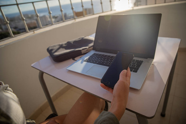 Chica sostiene su teléfono inteligente en la mano mientras trabaja con su computadora portátil sentado en el balcón. Nómada digital - Foto, imagen