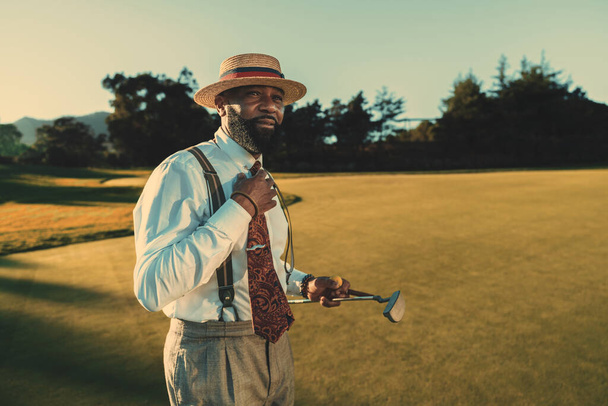 A portrait of a dapper mature black man in an elegant outfit with white a shirt, tie, and trousers with suspenders, standing on an evening golf field and holding his club and a ball; a copy space area - Fotó, kép