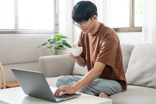 Technology Concept Mężczyzna w brązowej koszulce używając jednej ręki do pisania na laptopie, podczas gdy druga ręka trzyma filiżankę kawy. - Zdjęcie, obraz