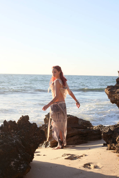  portret van mooi vrouwelijk schip vernield model dragen gescheurde jurk. poseren op de rotsachtige Oceaan kustlijn bij zonsondergang, - Foto, afbeelding