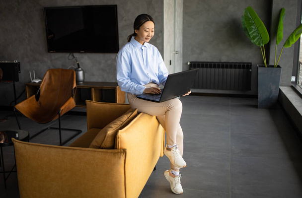 Donna d'affari asiatica che lavora con computer portatile seduto da solo su un divano giallo moderno in un interno dell'ufficio. - Foto, immagini