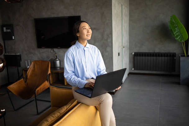 Azji businesswoman pracuje z laptopem siedzi sam na nowoczesnej, żółtej kanapie we wnętrzu biura. - Zdjęcie, obraz