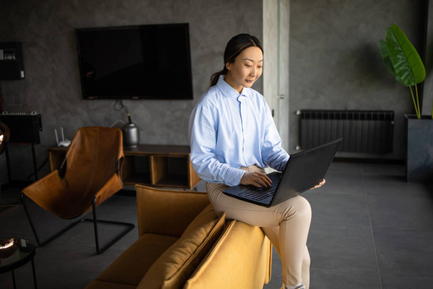 Donna d'affari asiatica che lavora con computer portatile seduto da solo su un divano giallo moderno in un interno dell'ufficio. - Foto, immagini