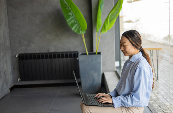 Azji businesswoman pracuje z laptopem siedzi sam na nowoczesnej, żółtej kanapie we wnętrzu biura. - Zdjęcie, obraz
