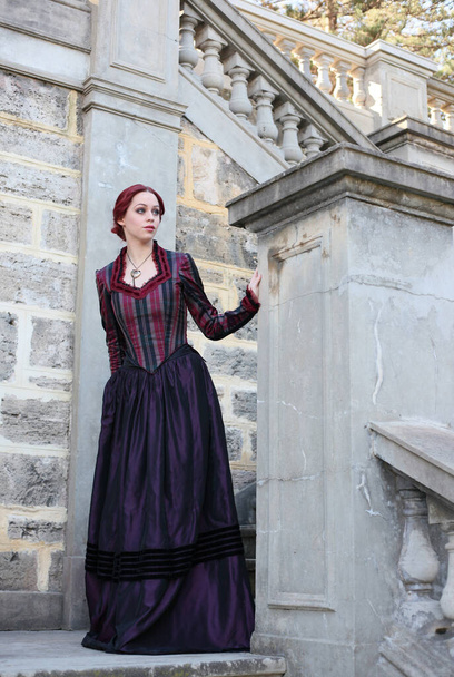 ritratto di donna dai capelli rossi che indossa uno storico costume da camice vittoriano, passeggiando per una splendida posizione con architettura in pietra. - Foto, immagini