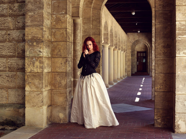 Full length portrait of red-haired woman wearing a  beautiful gothic gown costume, walking around  location with  romantic castle stone architecture background. - Photo, Image