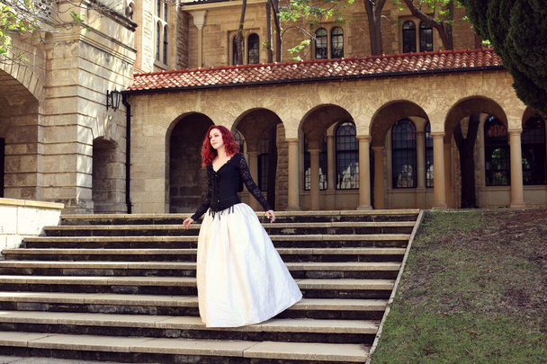 Teljes hosszúságú portré vörös hajú nő visel egy gyönyörű gótikus köntös jelmez, sétálni helyen romantikus várkő építészet háttér. - Fotó, kép
