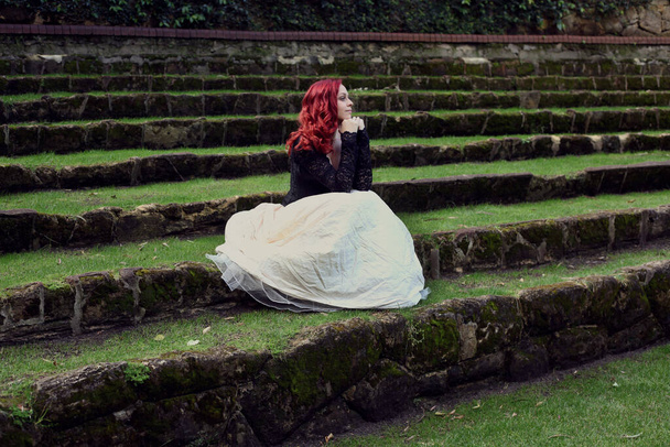 Полноразмерный портрет рыжеволосой женщины в красивом готическом костюме, прогуливающейся по местности на фоне романтической каменной архитектуры замка. - Фото, изображение