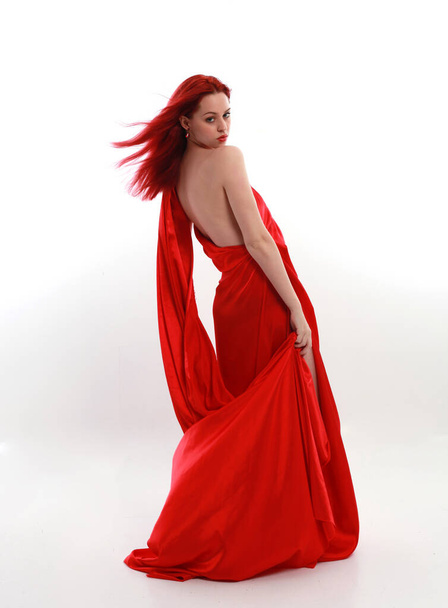 Teljes hosszúságú portré vörös hajú nő visel egy gyönyörű szexi selyem ruha, álló póz kreatív kar gesztusok, elszigetelt fehér stúdió háttér. - Fotó, kép