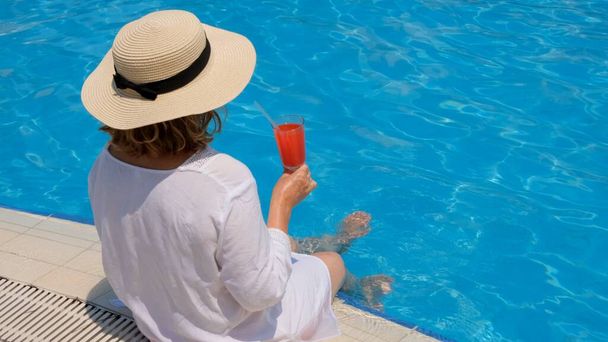 mulher em um chapéu de palha relaxa perto da piscina, bebe um coquetel rosa e balança as pernas na água. Conceito de humor verão - Foto, Imagem