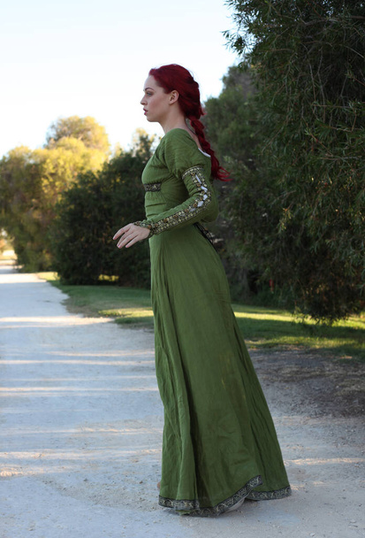 Πλήρες πορτρέτο της κοκκινομάλλας γυναίκας που φοράει ένα όμορφο πράσινο μεσαιωνικό φόρεμα φαντασίας. Τοποθέτηση με χειρονομιακά χέρια σε ένα μαγεμένο δάσος φόντο. - Φωτογραφία, εικόνα