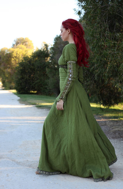 Ganzkörperporträt einer rothaarigen Frau in einem wunderschönen grünen mittelalterlichen Fantasiegewand. Posieren mit gestikulierenden Händen auf einem verzauberten Waldhintergrund. - Foto, Bild
