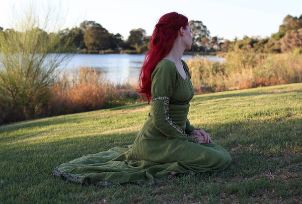 Plně dlouhý portrét rudovlasé ženy v krásných zelených středověkých fantasy šatech. Pózování s gesturálními rukama na začarovaném lesním pozadí. - Fotografie, Obrázek