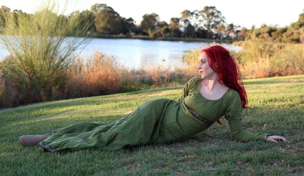 Ortaçağ fantezi elbisesi giymiş kızıl saçlı bir kadının tam boy portresi. Büyülü bir orman arka planında el hareketleriyle poz vermek. - Fotoğraf, Görsel