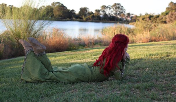 Portrait complet de femme aux cheveux roux portant une belle robe fantaisie médiévale verte. Poser avec des mains gestuelles sur un fond de forêt enchantée. - Photo, image
