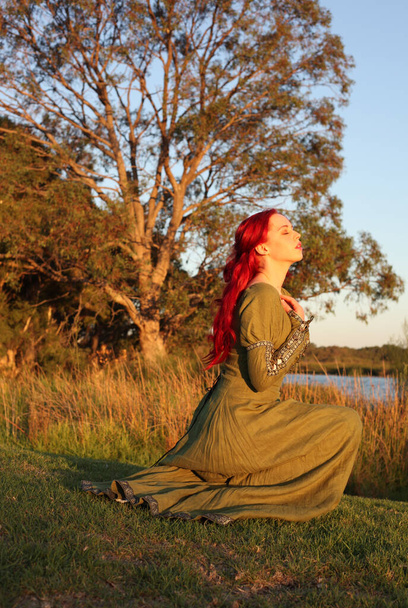 Ganzkörperporträt einer rothaarigen Frau in einem wunderschönen grünen mittelalterlichen Fantasiegewand. Posieren mit gestikulierenden Händen auf einem verzauberten Waldhintergrund. - Foto, Bild