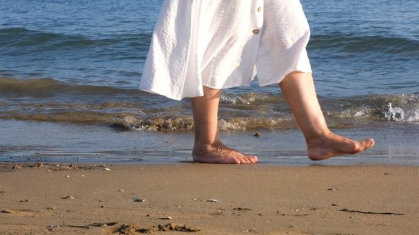 mature female legs are walking along the sandy beach and splashing in the sea on a summer sunny day. Woman walks barefoot on the water close-up - Photo, Image