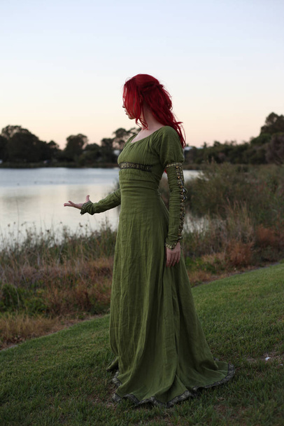 Ortaçağ fantezi elbisesi giymiş kızıl saçlı bir kadının tam boy portresi. Büyülü bir orman arka planında el hareketleriyle poz vermek. - Fotoğraf, Görsel
