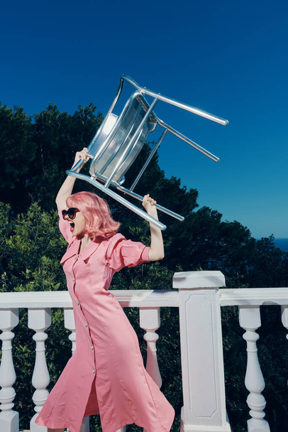 γυναίκα με ροζ μαλλιά σε γυαλιά με μια καρέκλα στα χέρια της στο μπαλκόνι - Φωτογραφία, εικόνα