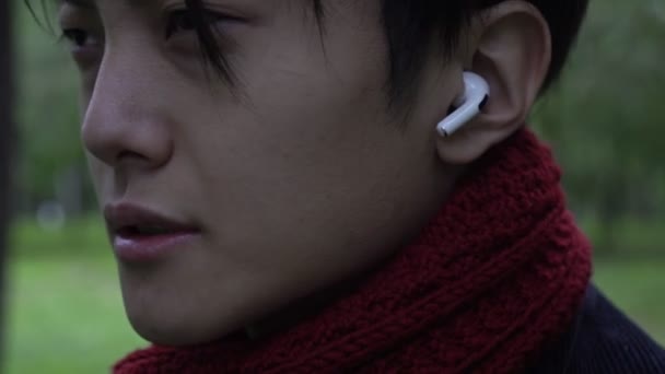 Un uomo ascolta musica con le cuffie senza fili. Giovane asiatico che indossa le cuffie all'aperto. Auricolare wireless nell'orecchio primo piano - Filmati, video