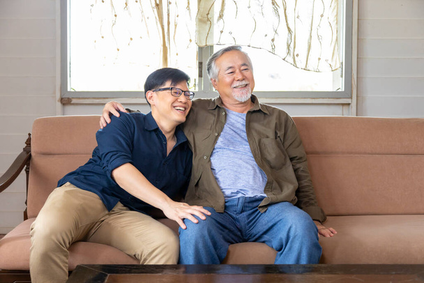 Старший азіатський чоловік і його син обіймають одне одного посміхаючись обличчям на дивані в його пенсійному домі. - Фото, зображення