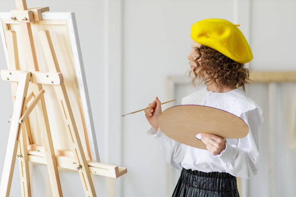 Portrait of little girl artist in yellow beret who paints picture in studio while holds pallete  - Foto, imagen