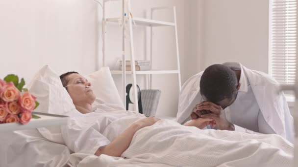 Mediana toma de triste hombre afroamericano cogido de la mano de su esposa enferma en coma acostada en la cama del hospital - Imágenes, Vídeo