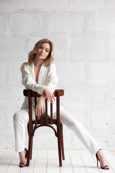 A girl in a white suit with loose dry hair and makeup is sitting on a chair on a white background. A fashionable full-length studio portrait. Elegant fashionable woman - Fotó, kép