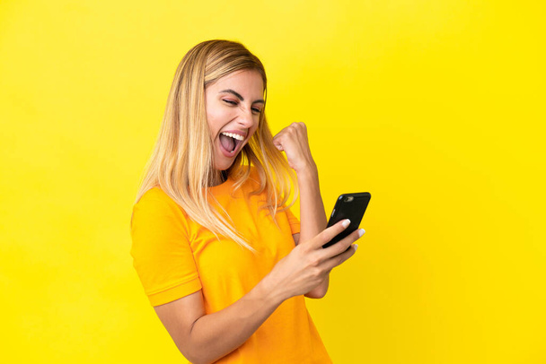 Blondi Uruguayn tyttö eristetty keltaisella taustalla puhelimen voittoasennossa - Valokuva, kuva