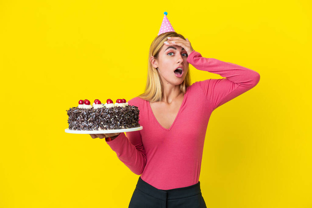 Blonde Uruguayenne fille tenant gâteau d'anniversaire isolé sur fond jaune faisant geste surprise tout en regardant sur le côté - Photo, image