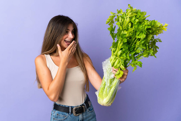 Young woman holding a celery isolated on purple background with surprise and shocked facial expression - Photo, Image