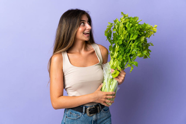 Young woman holding a celery isolated on purple background looking up while smiling - Фото, изображение