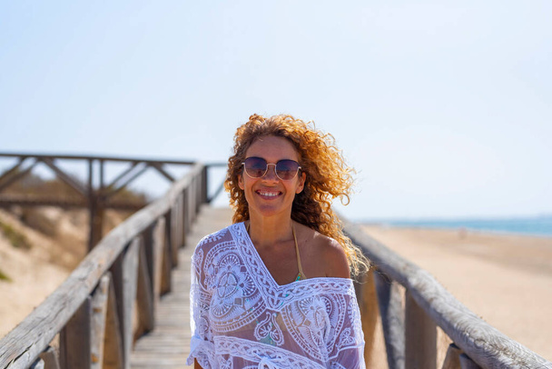 Portrét šťastné a veselé mladé dospělé ženy usmívající se na pláži. Sebevědomé turistické a letní cestování dovolená dovolená životní styl. Krásná dáma se slunečními brýlemi a bílými šaty venku - Fotografie, Obrázek