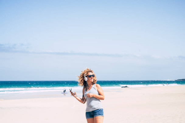 Feliz turista femenina disfrutar de ocio al aire libre en la playa escuchando música desde el teléfono móvil y auriculares amiling y disfrutar de vacaciones de verano estilo de vida de viajes. Mujer bonita alegre - Foto, Imagen